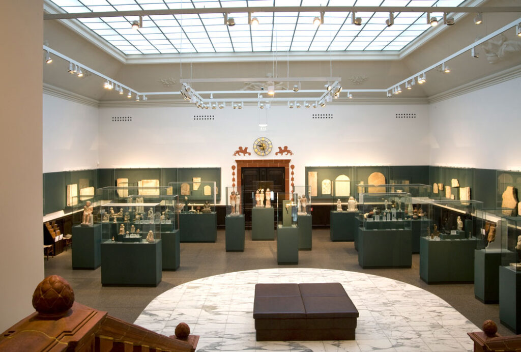Blick in einen hellen Ausstellungsraum des Ägyptischen Museums Leipzig mit Vitrinen und ägyptischen Artefakten auf Sockeln und an den Wänden sowie einer Sitzbank in der Raummitte
