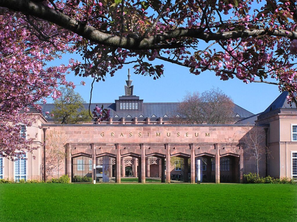 Das Grassimuseum mit blühenden Kirschbaumzweigen im Vordergrund und grünem Rasen davor.