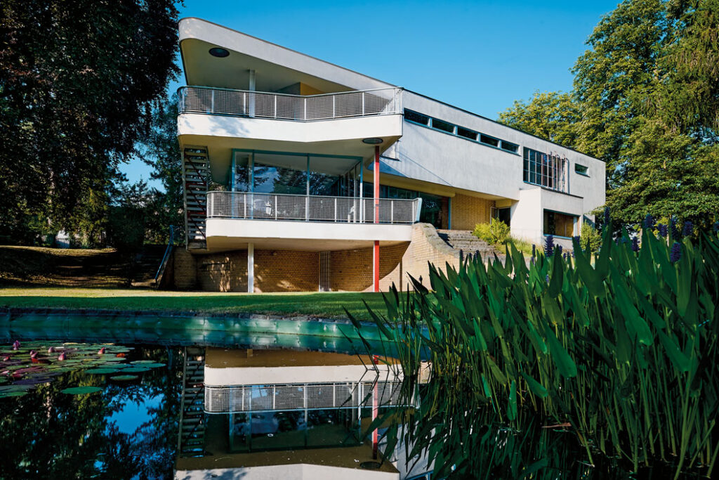 Modernes, kubisch gestaltetes Haus Schminke mit großen Fenstern, Balkonen und rotem Stützpfeiler, umgeben von Bäumen und mit Teich im Vordergrund, der das Gebäude spiegelt.
