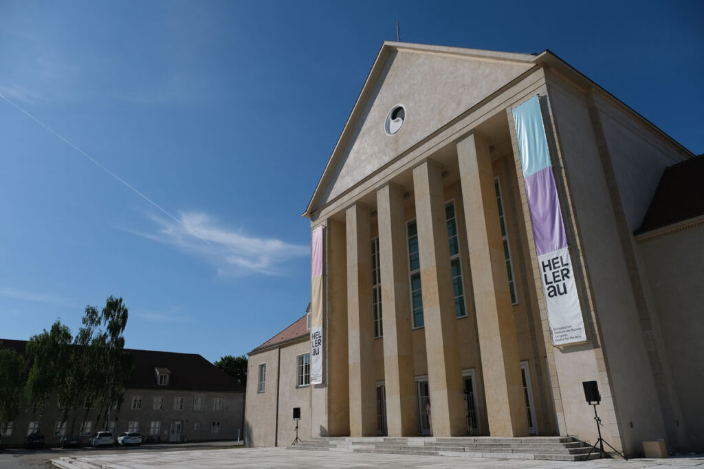Großes klassizistisches Gebäude mit hohen Säulen und zwei vertikalen Bannern mit der Aufschrift 'HELLERAU' vor blauem Himmel
