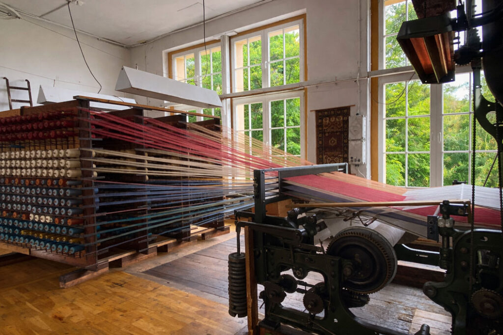 Webmaschine in einer Schauweberei mit vielen farbigen Garnspulen und Fäden, die durch den Webstuhl verlaufen.