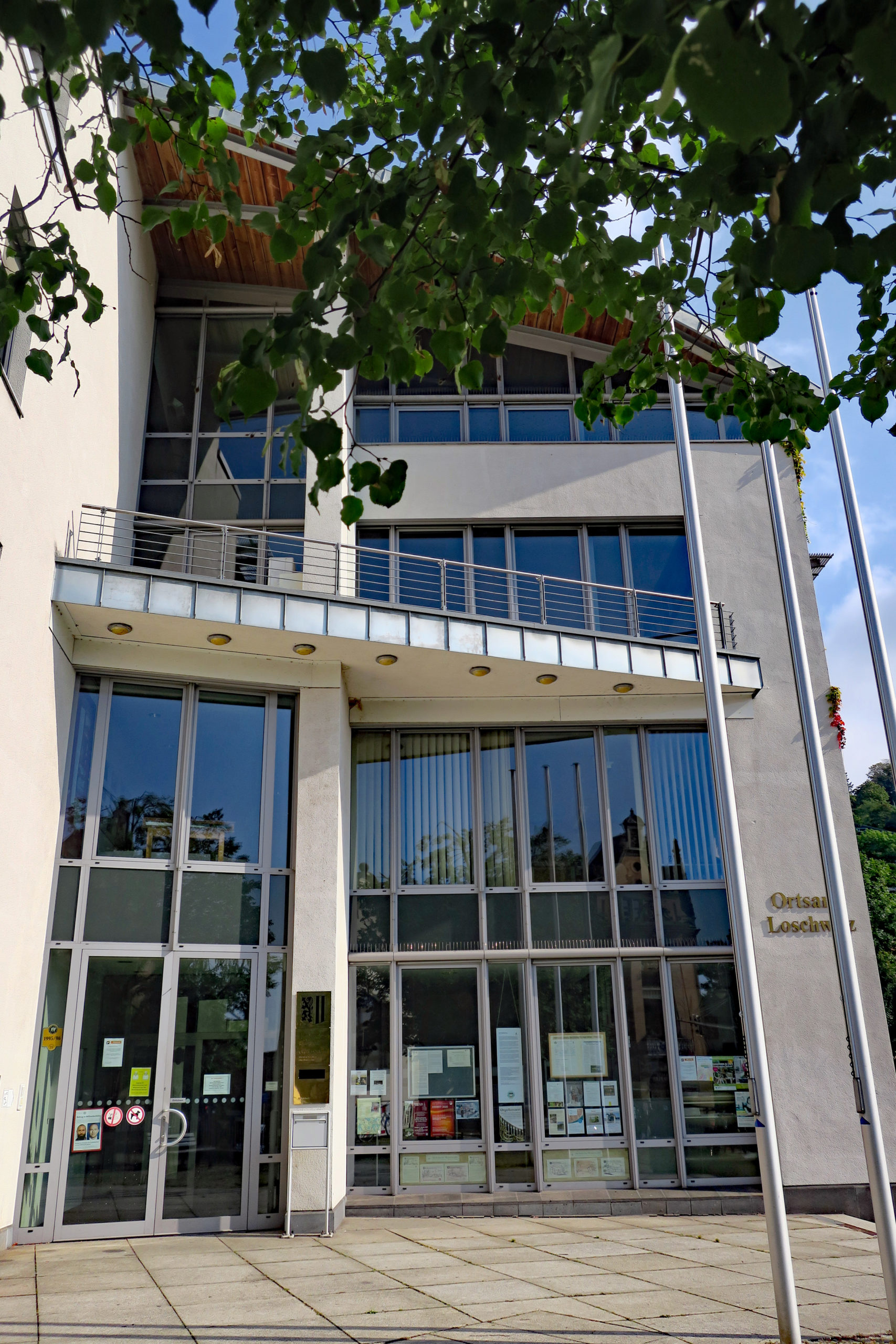 Das Rathaus Dresden Loschwitz ist geprägt von einer großen Glasfront, in der sich die gegenüberliegenden Gebäude und der blaue Himmel spiegeln. Auf dem Vorplatz sind quadratische Steinplatten verlegt, auf der rechten Seite stehen drei Fahnenmasten und vom oberen Bildrand ragen Lindenblätter in das Bild.