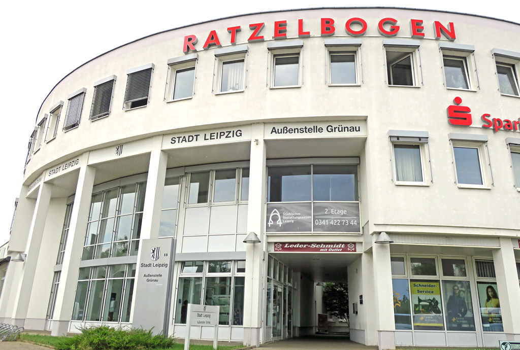 Rundes weißes Verwaltungsgebäude, drei Etagen und am obersten Gebäuderand in roten großen Buchstaben der Schriftzug "Ratzelbogen".