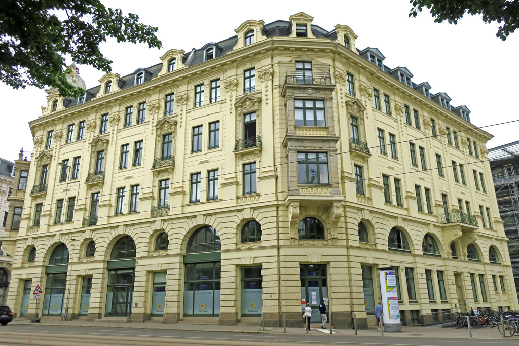 Das Gebäude, in dem sich das Bürgerbüro Otto-Schill-Straße befindet, nimmt fast das ganze Bild ein. Insgesamt hat das Gebäude fünf Etagen. In beiden oberen Stockwerken, direkt über den barrierefreien Haupteingang auf der Ecke, sind Erker. In der darauffolgenden Etage sind ein Balkon und darüber eine Fenstergaube. In der ersten Etage haben die Fenster und Eingangstüren oberhalb einen halbrunden Fensterbogen. Die Etagen zwei bis vier gibt es Fenster mit Verzierungen und zum Teil kleine Balkone. Die fünfte Etage hat komplett Gauben, als einfaches und Doppelfenster. Rechts um die Ecke ist der Zugang für Rollstuhlfahrer mit Rampe. Auf dem Vorplatz des Nebeneingangs steht eine Litfaßsäule und Fahrradständer mit angeschlossenen Rädern. Im Vordergrund befindet sich die Straße mit Straßenbahnschienen. Am oberen Rand ragen Äste mit grünen Blättern in das Bild.