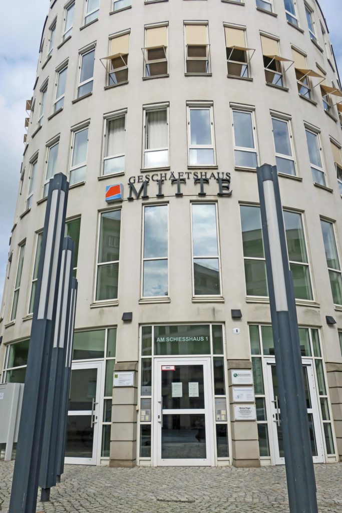 Halbrundes Geschäftshaus Dresden-Mitte, welches fünf Etagen mit länglich schmalen Fenstern und drei Glaseingangstüren hat. Der Vorplatz ist gepflastert und im Hintergrund ist blauer Himmel mit weißen Schleierwolken.