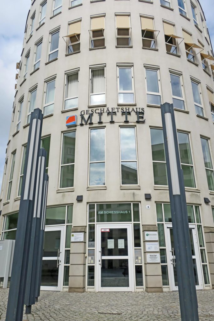 Halbrundes Geschäftshaus Dresden-Mitte, welches fünf Etagen mit länglich schmalen Fenstern und drei Glaseingangstüren hat. Der Vorplatz ist gepflastert  und im Hintergrund ist blauer Himmel mit weißen Schleierwolken.