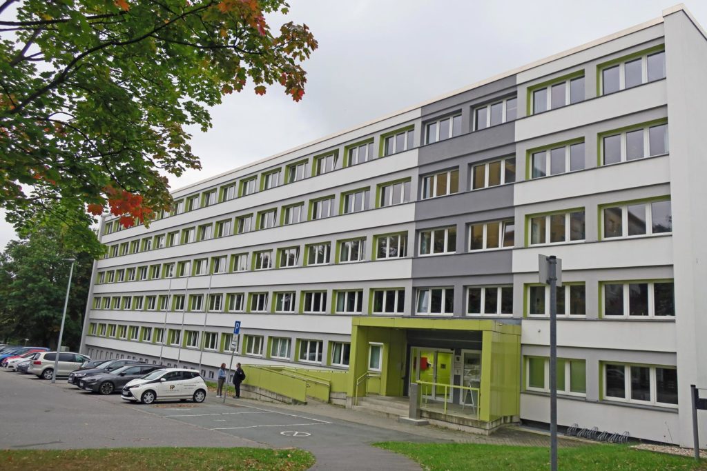 Schräger Blick auf das Finanzamt Süd, welches fünf Etagen hat, der Eingangsbereich hat einen grünen Anstrich und das Gebäude ist grau-weiß angestrichen. Rechts neben dem Eingang stehen Fahrradständer, links vor dem Gebäude stehen Autos auf dem Parkplatz, im Vordergrund ist Rasenfläche und vom oberen linken Rand ragen Äste eines herbstlich gefärbten Ahornbaumes in das Bild..