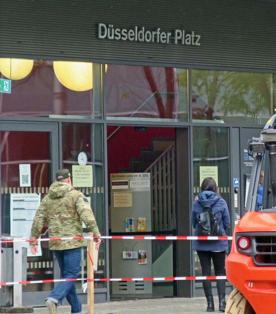 Eingangsbereich zur Bürgerservicestelle Am Wall von der Düsseldorfer Platz aus. Die Eingangstür aus Glas und in eine Glasfront integriert. Zwei Personen gehen darauf zu, um das Gebäude zu betreten. Über dem Eingangsbereich ist der Schrift "Düsseldorfer Platz". Im Vordergrund steht auf der rechten Seite ein Bagger und die Baustelle ist mit rot-weißem Absperrband gesichert.