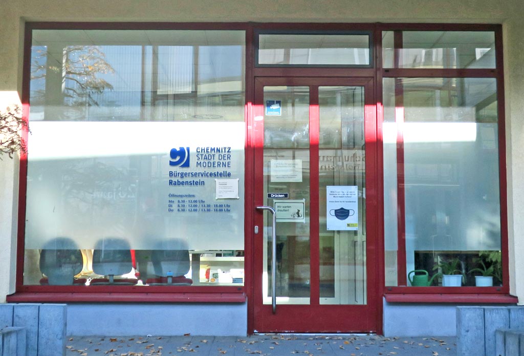 Der Eingang zur Bürgerservicestelle Rabenstein besteht aus einer großen Glasfront. Die Tür und die Fenster haben eine dunkelrote Umrandung. Auf den Glasscheiben ist ein breiter Milchglasstreifen und in dunkelblauer Schrift sind die Öffnungszeiten abgebildet. Hinter den Glasscheiben stehen auf der linken Seite drei Stühle im Warteraum und auf der rechten Seite zwei Pflanzen bzw. eine Gießkanne.