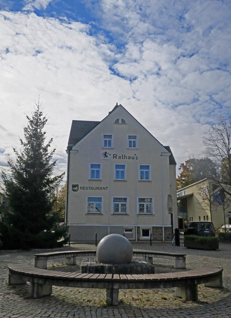 Das Rathaus in Röhrsdorf ist dreietagig, hat einen Sandsteinsockel, ein hellgelben Anstrich und in dunkelgrün die Schriftzüge "Rathaus" und "Restaurant". Link neben dem Gebäude steht ein großer Nadelbaum und auf der rechten Seite ist das Gebäude der Kindertageseinrichtung bzw. ein parkendes Auto. Auf dem gepflastertem Vorplatz steht eine Sitzgruppe um einen modernen Springbrunnen.