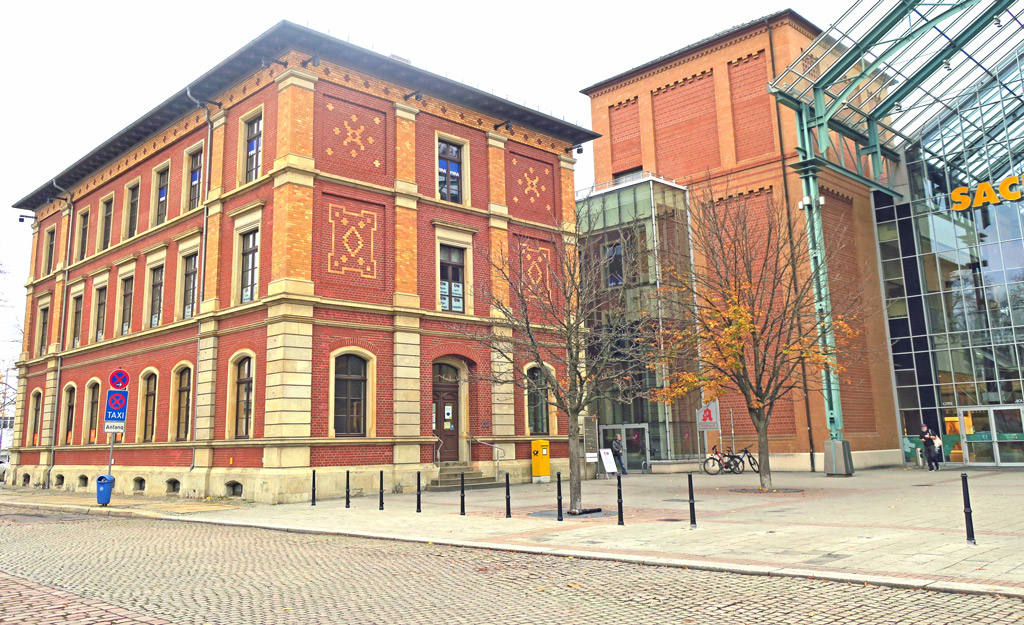 Auf der linken Hälfte ist ein dreistöckiges rotes Backsteinhaus. In der untersten Etage befindet sich die Bürgerservicestelle. Auf der rechten Seite ist ein Teil des Sachsenallee-Centers zuerkennen, welcher ebenfalls aus Backstein und einer großen Glasfront besteht. Auf dem gepflastertem Vorplatz stehen Bäume, Fahrräder und Poller.