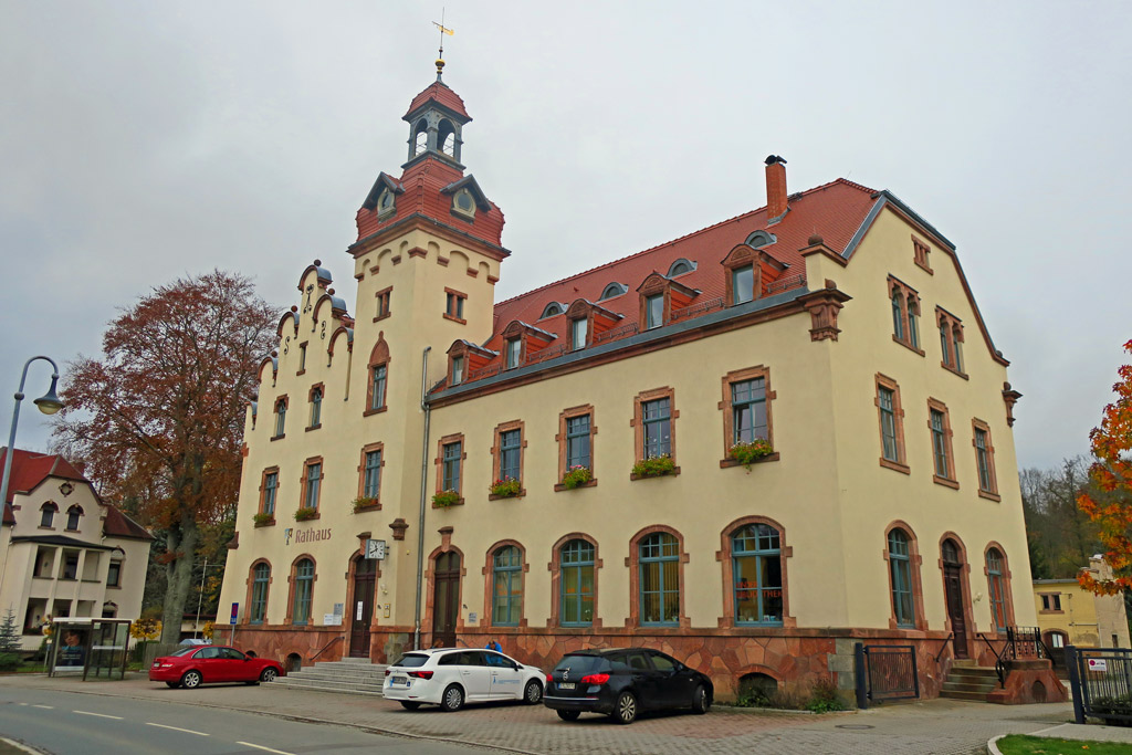 Rathausgebäude von Einsiedel in Vollansicht. Das Gebäude hat drei Etagen, eine Treppe zu zwei Eingangstüren und dazwischen hängt eine Uhr, einen seitlichen Eingang und einen Rathausturm mit Weiterfahne. Vor dem Gebäude stehen drei Autos.