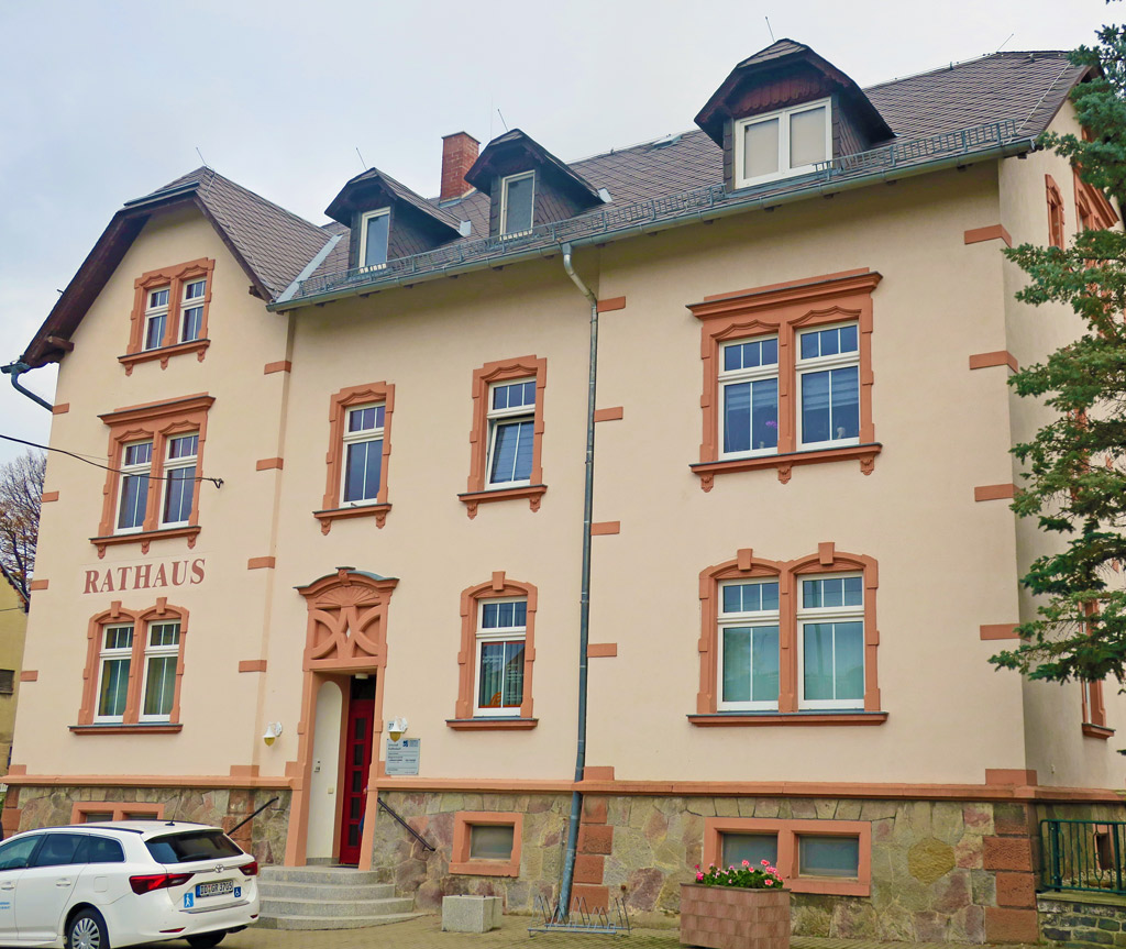 Das Rathaus Klaffenbach hat drei Etagen und ein Dachgeschoss. In das Gebäude führt eine Treppe mit vier Stufen zu einer dunkelroten Eingangstür, die Fassade hat einen hellrosa Anstrich, die Fenster sind weiß, mit einer hellroten Fensterlaibung und das ist mit schwarzem Schiefer gedeckt. Am rechten Bildrand ragen Zweige eines Baumes hinein. Direkt vor dem Gebäude parkt ein Auto.