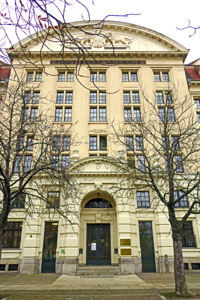 Im Hintergrund ist der Haupteingang des Finanzamtes Leipzig. Das Gebäude hat sieben Etagen. Auf dem Vorplatz sind große und kleine Pflastersteine sowie große Gehwegplatten verlegt. Im Vordergrund stehen zwei Bäume und von oben ragen Äste in das Bild. Die Bäume und Äste haben kein Laub, da die Aufnahme des Bildes im Spätherbst erfolgte.