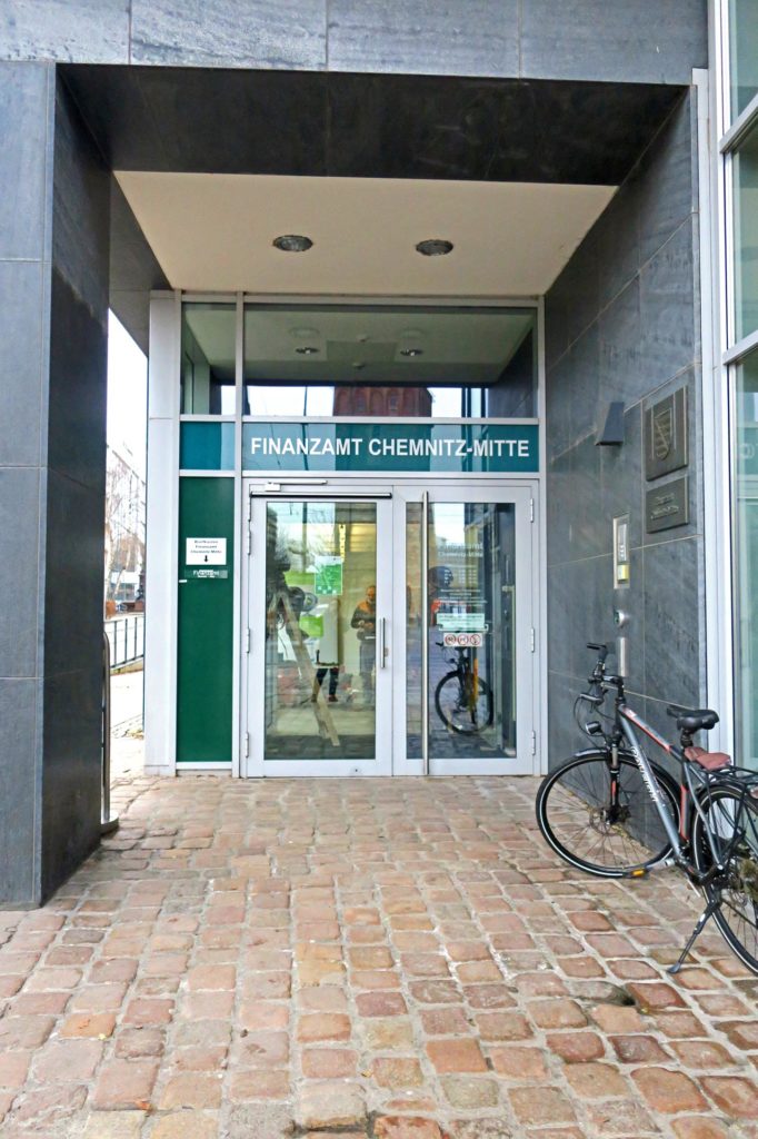 Eingangsbereich Finanzamt Mitte. Die Glastür zum Amt hat eine weiße Umrandung und oberhalb steht der Schriftzug in weiß "Finanzamt Chemnitz-Mitte" auf grünem Glas. Der Weg davor ist mit roten Ziegelsteinen gepflastert, auf der linken Seite steht ein Fahrrad und links eine dunkelgraue Säule.