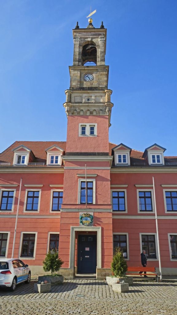Das Rathaus hat drei Etagen, einen roten Farbanstrich und einen Rathausturm mit goldener Wetterfahne. Auf dem Vorplatz steht links ein Auto, mittig Pflanzkübel aus Stein und auf der rechten Seite eine mahagonifarbene Holzbank. Im Hintergrund ist blauer Himmel.