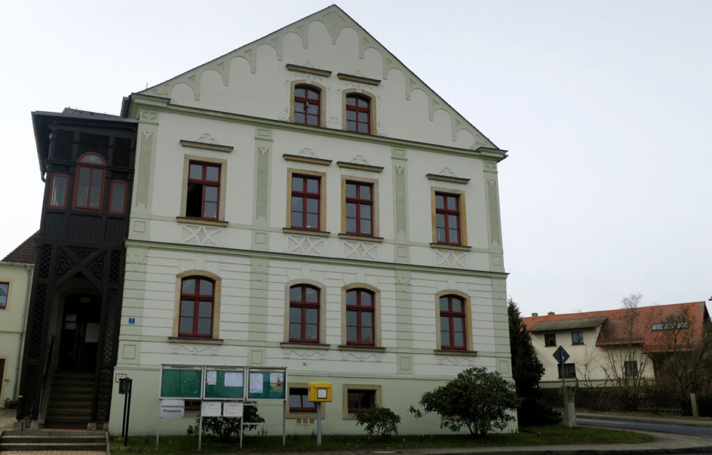 Das Gebäude hat drei Etagen und einen Fachwerkanbau aus dunklem Holz. Vor der Gemeindeverwaltung stehen auf einer Rasenfläche kleine Sträucher, eine Anzeigetafel und ein Briefkasten. Auf der rechten Seite neben dem Gebäude stehen ein Baum und ein Einfamilienhaus. Der Himmel im Hintergrund ist bedeckt.