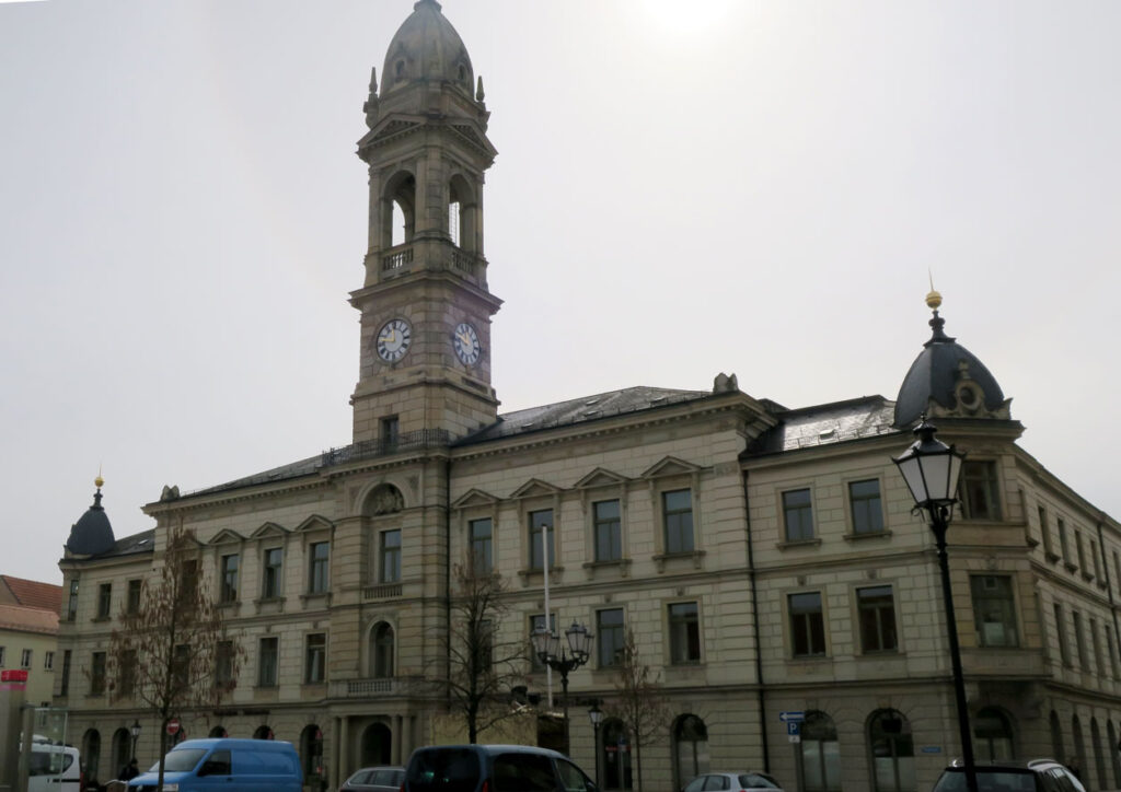 Das Rathaus hat drei Etagen, auf der linken und rechten Seite ein kleines Türmchen und über dem Eingang einen großen Rathausturm mit Uhr. Auf dem Marktplatz vor dem Gebäude parken Autos, und der Himmel im Hintergrund ist sehr stark bedeckt.