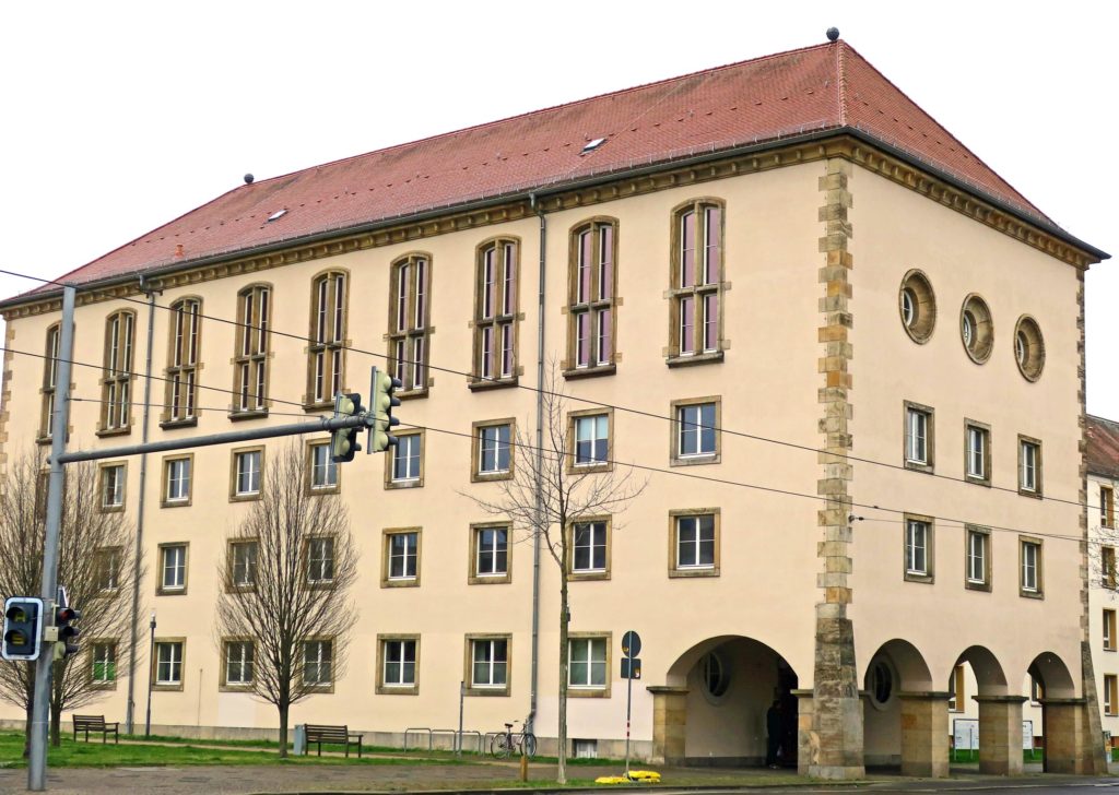 Gebäude mit vier Etagen, einem Durchgang mit Rundbögen und einem roten Ziegeldach. In der untersten Etage ist das Bürgerbüro. Vor dem Gebäude auf der linken Seite stehen drei Bäume auf einer Wiese und auf dem Gehweg steht eine Ampel, welche in das Bild ragt.