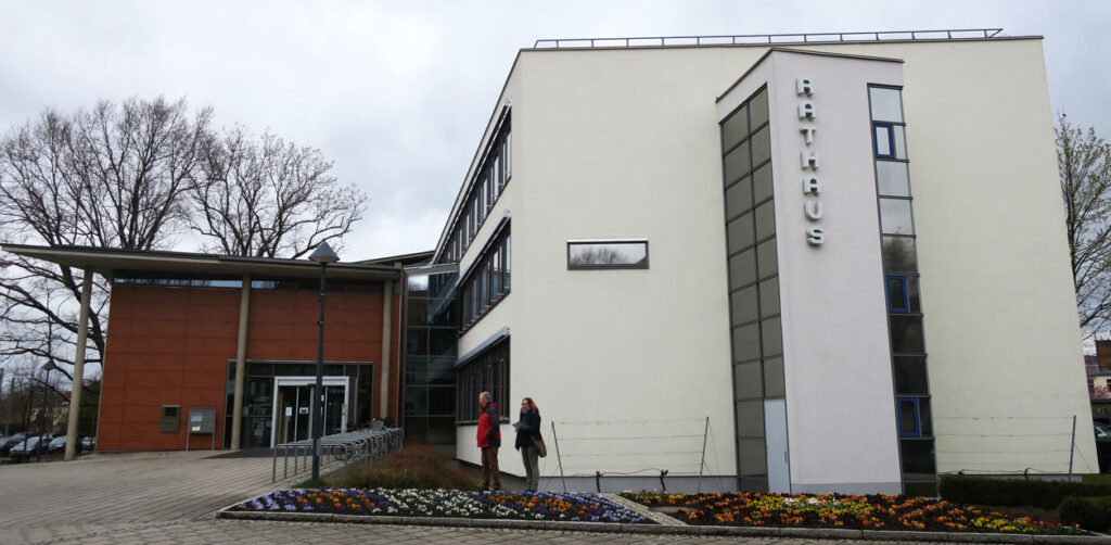 Das Verwaltungsgebäude ist ein moderner Neubau mit drei Etagen. Die Fassade ist weiß, der Eingangsbereich terracottafarben und mit einer großen Glastür versehen. Vor dem Eingang gibt es rechts Fahrradständer, in den Beeten vor dem Hauptgebäude sind Frühblüher gepflanzt. Im Hintergrund stehen links und rechts Bäume, und der Himmel ist bedeckt.