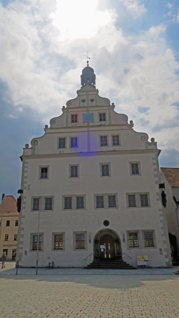 Das Gebäude hat drei Etagen sowie drei Dachetagen und Rathausturm mit Wetterfahne. Die Tür- und Fenstereinfassungen sind aus Sandstein, die Fassade hat einen eierschalenfarbenen Anstrich, im zweiten Dachgeschoss ist zwischen den beiden Fenstern eine quadratische Rathausuhr mit blauem Untergrund sowie goldenen Ziffernblatt und Zeigern. Der Marktplatz ist gepflastert, im Hintergrund sind Wohnhäuser und der Himmel ist blau, mit Schleierwolken, durch die die Sonne strahlt.