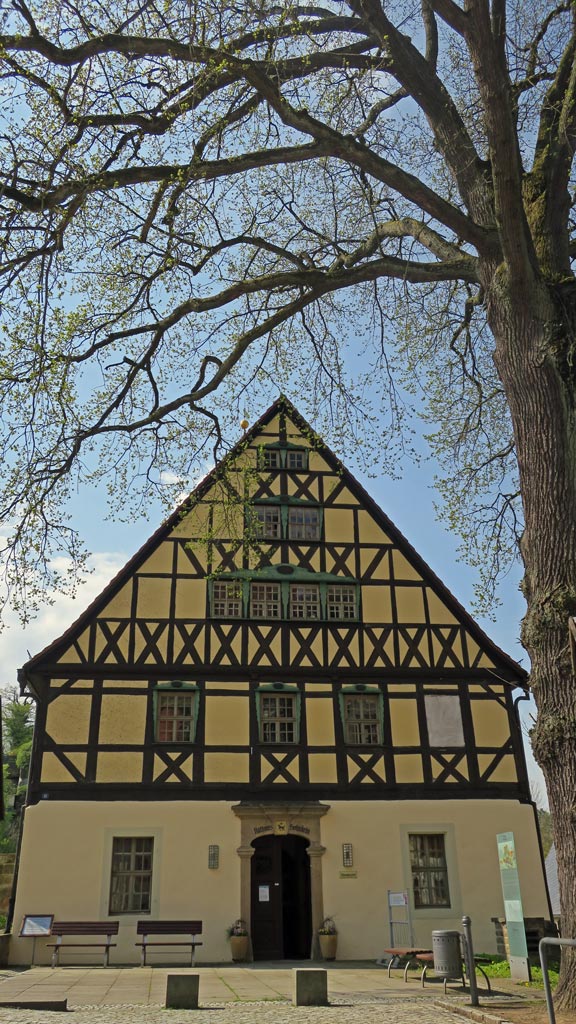 Das Gebäude hat zwei Etagen sowie drei Dachetagen. Es handelt sich um ein Umgebindehaus. Die unterste Etage hat einen sandfarbenen und die restlichen Etagen einen gelben Anstrich. Die Fensterrahmen sind grün und die Eingangstür dunkelbraun. Auf dem Vorplatz stehen Bänke, Hinweistafeln, ein Abfalleimer sowie ein Fahrradständer rechts neben der Eingangstür. Am rechten Bildrand steht eine große Linder, deren Äste von oben in das Bild ragen. Der Himmel im Hintergrund ist blau, mit kleinen Schleierwolken.