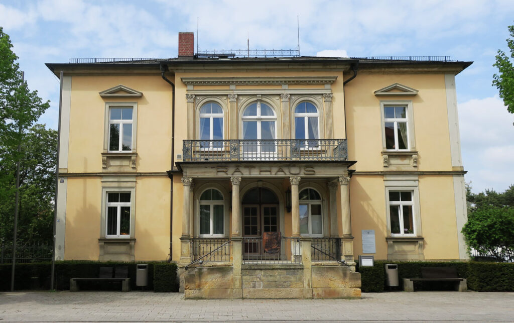 Das Rathaus hat zwei Etagen, eine pastellgelbe Fassade und eine Sandsteintreppe, die zum Eingang führt. Auf der linken und rechten Seite vor dem Gebäude gibt es eine kurzgehaltene Hecke. Davor stehen auf beiden Seiten eine Bank und ein Mülleimer. Der Fußweg ist gepflastert, neben dem Rathaus stehen Bäume, und der Himmel im Hintergrund ist blau mit großen Schäfchenwolken.