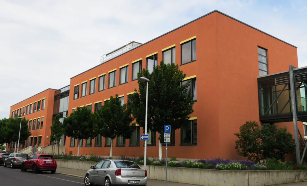 Schräge Ansicht auf das Landratsamt von rechts nach links. Das Gebäude hat drei Etagen, eine terrakottafarbene Fassade und der Eingangsbereich ist eine große Glasfront. Vor dem Gebäude stehen rechts und links neben dem Eingangsbereich Bäume und darunter sind Bodendecker gepflanzt. Auf dem Gehweg stehen Laternen und Verkehrsschilder. Auf der Straße parken Autos und der Himmel im Hintergrund ist bedeckt.