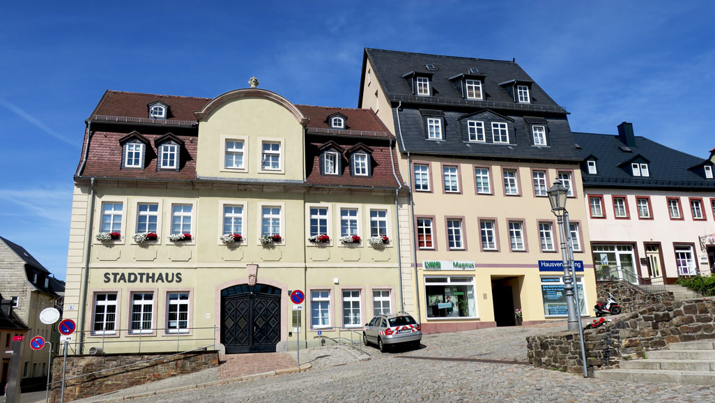 Ansicht auf drei aneinander gereihte Fachwerkhäuser. Im ersten Haus ganz links ist das Stadthaus inklusive Bürgerbüro. Dieses hat drei Etagen. In der zweiten Etage stehen Blumenkästen auf den Fensterbänken. Es gibt einen barrierefreien Parkplatz auf der rechten Seite vor der Eingangstür. Der Weg und die Straße davor sind gepflastert. Der Himmel im Hintergrund ist blau, mit kleinen Schleierwolken.