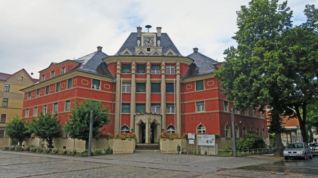 Das Rathaus ist ein Eckgebäude, hat vier Etagen, die Fassade ist dunkelrot sowie Sandsteinsäulen, das Dach ist mit Schiefer gedeckt und über der vierten Etage gibt es mittig eine Rathausuhr. Davor ist der Marktplatz mit drei Fahrradständern aus Edelstahl, links und rechts stehen jeweils eine moderne Straßenlaterne. Am rechten Bildrand parken Autos und auf der linken Seite steht ein Mehrfamilienhaus. Der Hintergrund ist bewölkt.