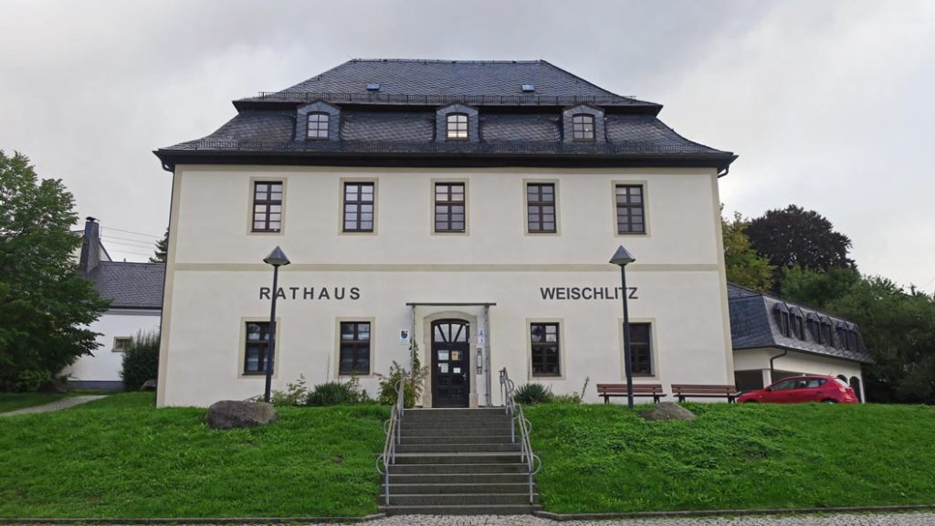 Das Gebäude hat zwei Etagen sowie ein Dachgeschoss, die Fassade ist sandfarben gestrichen, die Eingangstür sowie die Fensterrahmen sind dunkelbraun und das Dach ist mit Schiefer gedeckt. Mittig führt eine Treppe von der unteren Parkebene direkt zum Haupteingang, welcher ebenfalls in der Mitte des Gebäudes ist. Links und rechts von dem Treppenaufgang sind Grasflächen. Auf der rechten Seite vor dem Gebäude stehen zwei Bänke und auf der oberen Ebene parkt ein rotes Auto. Der Himmel im Hintergrund ist bedeckt.