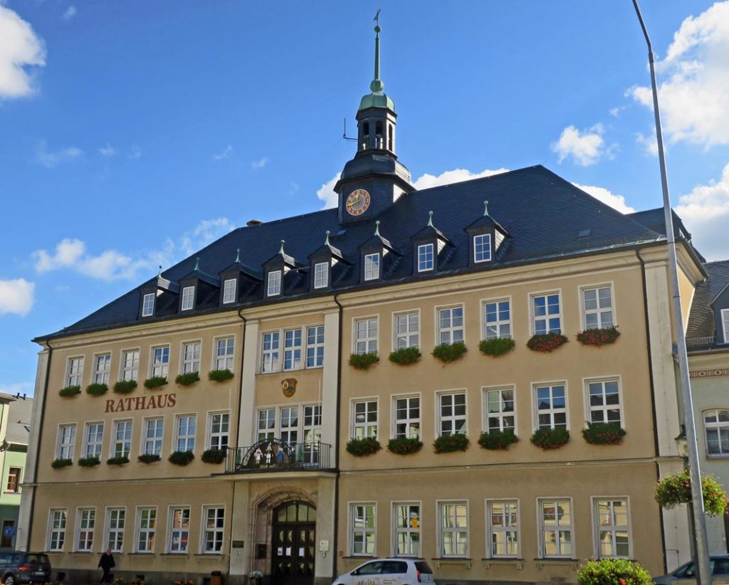 Die Stadtverwaltung Ehrenfriedersdorf hat drei Etagen und Dachgeschoss, welches mit Schiefer gedeckt ist, sieben Gauben und einen kleinen Rathausturm mit Uhr hat. Vor dem Gebäude stehen zwei Autos und auf dem Gehweg läuft eine Person. Im Hintergrund ist blauer Himmel mit weißen Wolken.