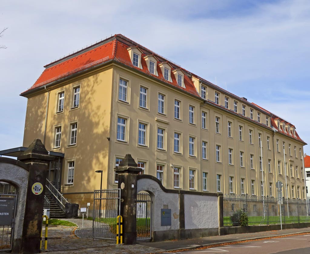 Schräge Ansicht auf das Finanzamtsgebäude der Stadt Freiberg. Dieses hat ein Keller, drei Etagen und ein Dachgeschoss. Der Farbanstrich ist ocker, das Dach mit rotem Biberschwanz gedeckt und Sockel ist aus Sandstein. Der Eingang ist auf der linken Seite des Gebäudes, zu diesem führt schräg ein gepflasterter Weg. Davor ist eine grüne Rasenfläche mit drei Fahnenmasten. Das Gelände ist umzäunt und am unteren Bildrand ist ein Stück Gehweg und Straße.