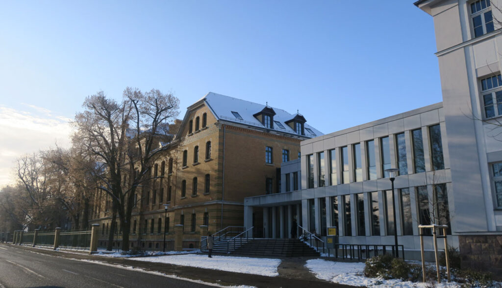 Schräge Ansicht auf das Finanzamt von rechts nach links. Das Gebäude hat links einen dreietagigen gelben Klinkerbau sowie ein Dachgeschoss, rechts davon ist ein moderner Bau über zwei Etagen. Dem schließt sich ein Altbau über drei Etagen mit einem Sandsteinsockel und einer grau-weißen Fassade an. Alle Gebäudeteile sind miteinander verbunden. Vor dem Gebäude liegt Schnee, es stehen Bäume und der Himmel im Hintergrund ist leicht bedeckt.