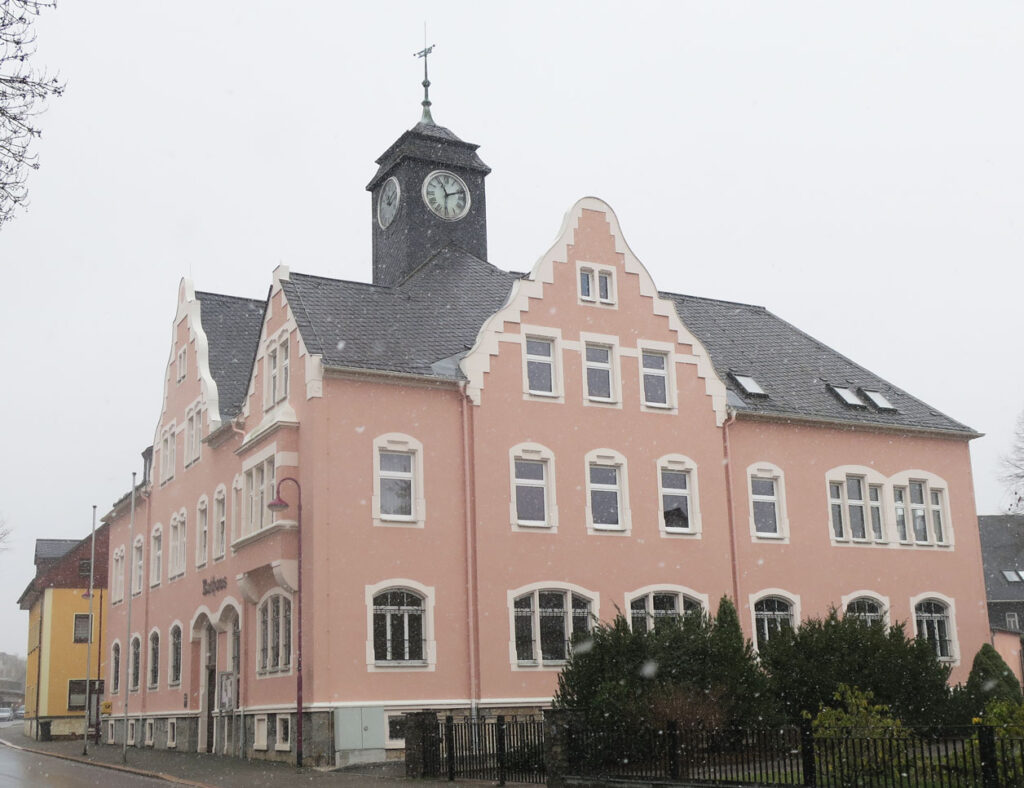 Schräge Ansicht auf die Gemeindeverwaltung, welche drei Etagen, eine altrosa Fassade, ein Schieferdach, weiße Tür- und Fensterleibungen und einen Turm mit Wetterfahne sowie Uhr hat. Am rechten Bildrand stehen vor dem Gebäude Büsche und links dahinter ein gelbes Einfamilienhaus. Der Himmel ist bedeckt und es schneit.