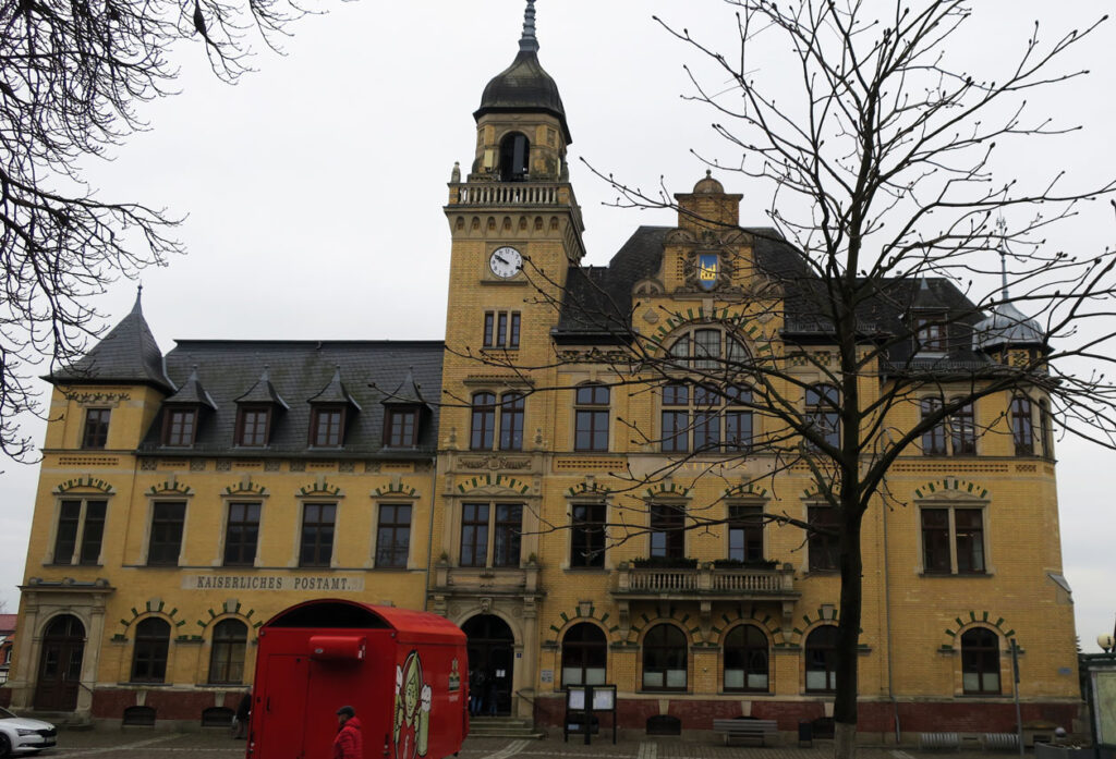Das Verwaltungsgebäude hat drei Etagen, der Sockel ist mit roten und die Fassade mit gelben Steinen verklinkert. Außerdem gibt es drei Türmchen, Gauben, eine Rathausuhr, einen Balkon und das Dach ist mit Schiefer gedeckt. Auf dem Vorplatz parkt ein weißes Auto, ein roter Bierwagen von Feldschlösschen. Rechts neben der Eingangstreppe stehen eine Anzeigetafel, eine Bank, ein Baum sowie ein Fahrradständer. Vom linken Bildrand ragen Zweige hinein. Der Himmel im Hintergrund ist bedeckt.