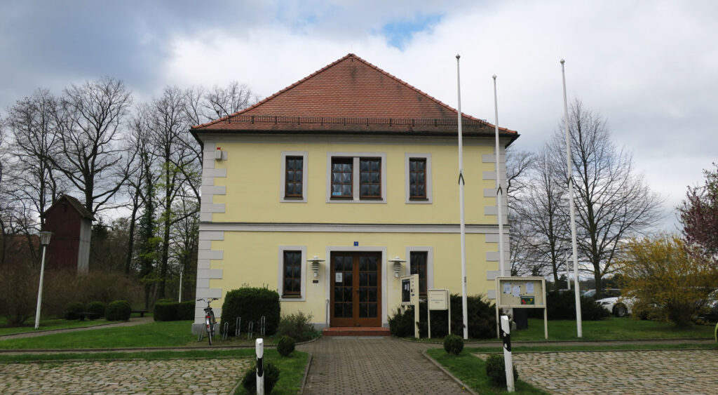 Ansicht auf das Verwaltungsgebäude, welches zwei Etagen, eine pastellgelbe Fassade und ein rotes Dach hat. Auf der linken Seite vor dem Gebäude sind kleine Sträucher und ein Fahrradständer mit einem Rad. Rechts neben dem Eingang sind kleine Sträucher, zwei Informationskästen, ein Briefkasten und drei Fahnenmasten. Auf beiden Seiten des Gebäudes sind Grünflächen mit Bäumen und großen Sträuchern. Parkflächen sind vor dem Gebäude links und rechts neben dem Weg sowie rechts neben dem Gebäude.