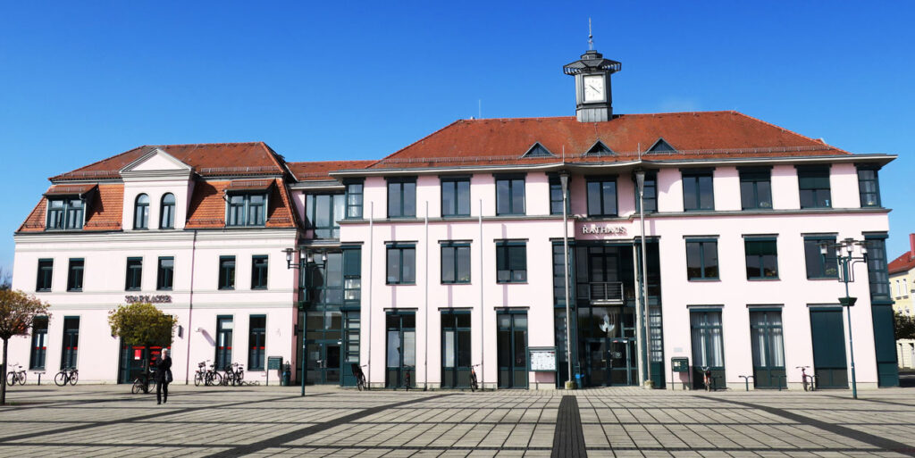 Ansicht vom Marktplatz auf das Rathaus, welches drei Etagen, ein rotes Dach und einen Rathausturm hat. Auf dem Marktplatz stehen Laternen mit Pflanzen und Fahnenmasten. Der Himmel im Hintergrund ist strahlend blau.