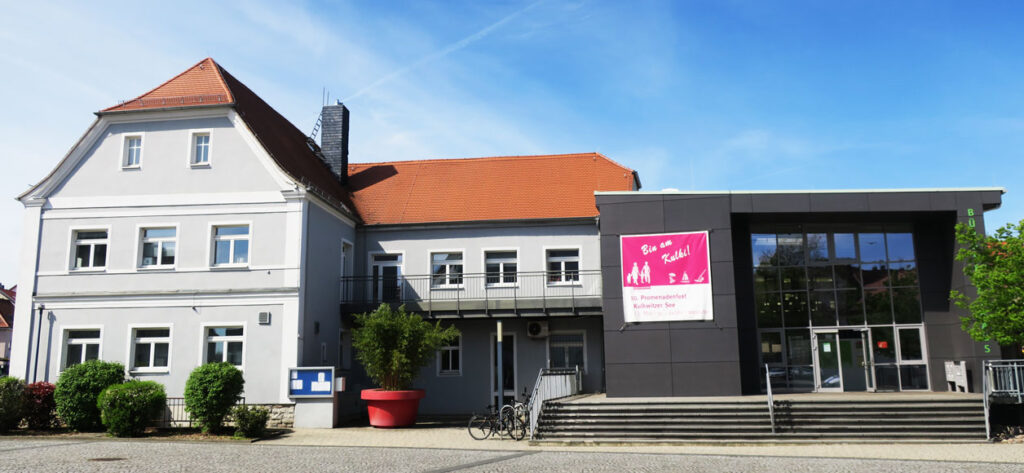 Das Verwaltungsgebäude hat links einen Altbau über zwei Etagen, ein Dachgeschoss, die Fassade ist hellgrau, und rechts einen modernen Neuanbau mit dunkelgrauer Fassade sowie einer großen Glasfront. Zum Neuanbau führt eine sehr breite Treppe mit einem Geländer links, mittig und rechts. Vor dem Gebäude sind auf der linken Seite Sträucher gepflanzt, es gibt eine Anzeigetafel und Fahrradständer. Der Vorplatz ist gepflastert und der Himmel im Hintergrund ist blau, mit großen weißen Schleierwolken.