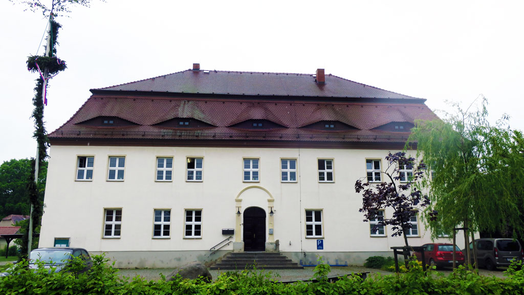Das Gebäude hat zwei Etagen, ein Dachgeschoss, die Fassade ist sandfarben, das Dach ist mit dunkelrotem Biberschwanz gedeckt und hat zwei Essen. Davor parken drei Autos, links steht ein Maibaum, rechts zwei kleine Laubbäume und am unteren Bildrand ist eine grüne Buchenhecke. Der Himmel im Hintergrund ist bedeckt.
