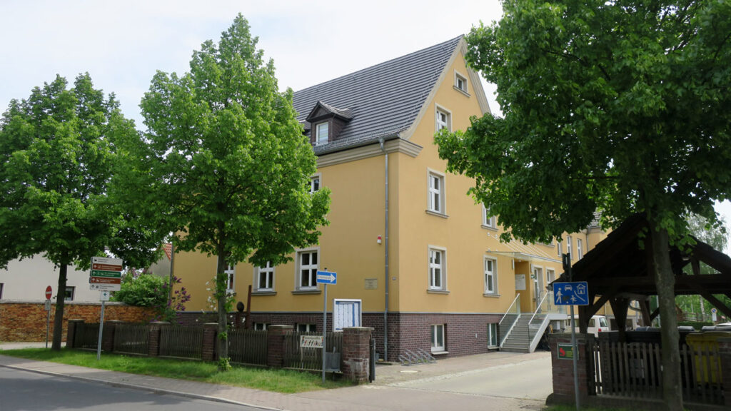 Schräge Ansicht auf das Gelände der Gemeindeverwaltung Schleife. Das Gebäude hat drei Etagen, ein Backsteinsockel, die Fassade ist gelb und das Dach ist mit Biberschwanz gedeckt. Vor dem Gelände stehen links neben der Einfahrt zwei Bäume auf einer Rassenfläche und links einer. Der Himmel im Hintergrund ist leicht bedeckt.