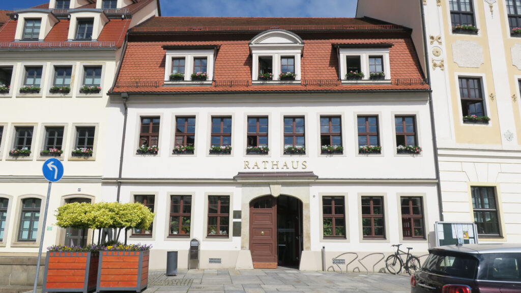 Frontansicht des Rathauses, welches drei Etagen, einen weißen Anstrich und ein rotes Biberschwanzdach hat. Jeweils auf der linken und rechten Seite stehen Stadthäuser. Vor dem Gebäude steht links ein Verkehrsschild, rechts daneben zwei große Pflanzenkübel mit kleinen Bäumen und Blumen. Direkt neben der Eingangstür ist links ein Mülleimer sowie ein Briefkasten, rechts Fahrradständer mit einem Rad und daneben eine Anzeigetafel mit Haushängen der Stadtverwaltung. Der Weg ist mit großen Platten ausgelegt, und im Hintergrund ist ein blauer Himmel mit Schleierwolken.