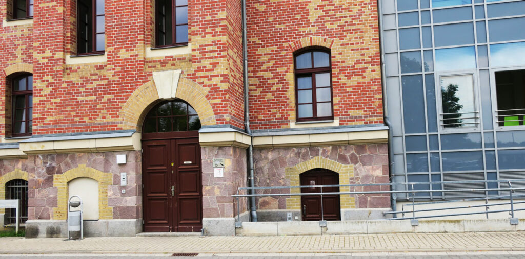 Es ist der barrierefreie Zugang des Landratsamtes Borna abgebildet. Auf der linken Seite ist ein Teil des Backsteingebäudes und rechts ein moderner Anbau aus Glas. Die Türen und Fenster sind braun. Auf dem Gehweg davor steht links ein Mülleimer.