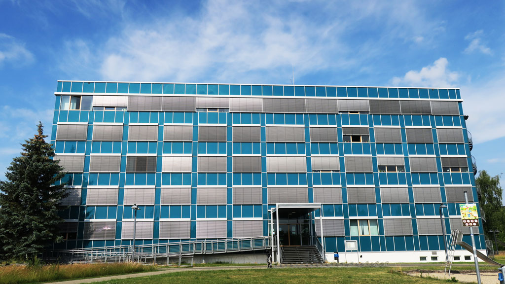 Das Gebäude ist ein Neubau, hat fünf Etagen, die Fassade hat eine dunkelblaue Verkleidung und an den Fenstern sind silbergraue Außenjalousien angebracht. Davor auf der linken Seite steht ein Nadelbaum. Auf dem Gehweg vor dem Eingang stehen zwei Laternen. Am unteren Bildrand ist eine Rasenfläche mit einem schräg betonierten Gehweg. Auf der rechten Seite am unteren Bildrand ist ein Spielplatz. Der Himmel im Hintergrund ist blau und hat weiße Schleierwolken.