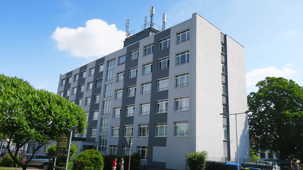 Das Gebäude hat fünf Etagen und ein Kellergeschoss. Die Fassade hat einen hell- und dunkelgrauen Anstrich. Am linken und rechten Bildrand stehen große Bäume. Der Himmel im Hintergrund ist blau und hat große weiße Wolken.