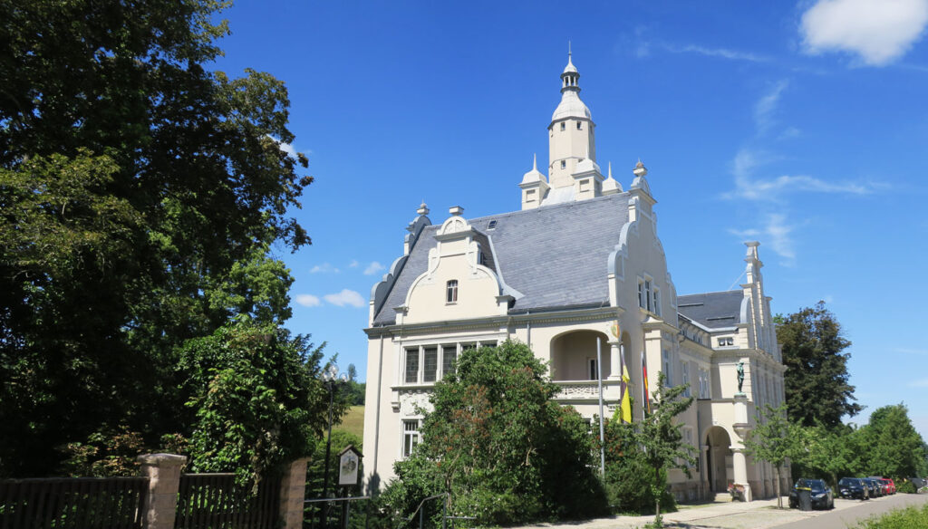 Schräge Ansicht auf die Stadtverwaltung Eibenstock. Auf der linken Seite stehen Bäume, rechts daneben das Rathaus und dann wieder Bäume. Das Ratshaus ähnelt einem Märchenschloss, welches viele Verzierungen, Türme und ein Schieferdach hat. Vor dem Rathaus gibt es zwei Fahnenmasten, und in den Parkbuchten stehen Autos. Der Hintergrund ist blau, mit vereinzelten weißen Wolken.