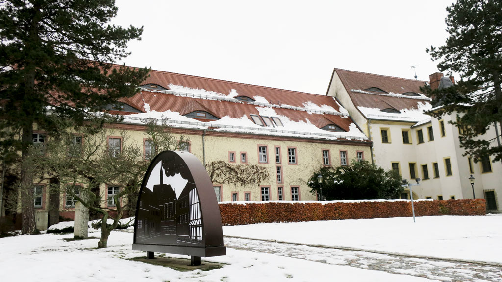 Ansicht auf einen Teil des Verwaltungsgebäudes, welches drei Etagen, Dachfenster und Gauben hat. Davor stehen links und rechts Bäume, eine Hecke und auf der Wiese ein großer Schwippbogen aus Metall. Der Himmel im Hintergrund ist bedeckt.