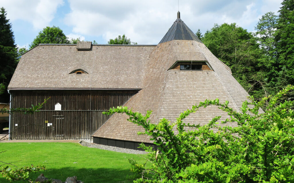 Pferdegöpel - Großes, historisch wirkendes Holzgebäude mit breitem, schindelgedecktem Dach. Rechts schließt sich ein runder, turmartiger Baukörper mit kegelförmigem Dach an. Das Gebäude ist von grünen Bäumen umgeben, davor liegt eine gepflegte Rasenfläche mit Sträuchern. Der Himmel ist leicht bewölkt.