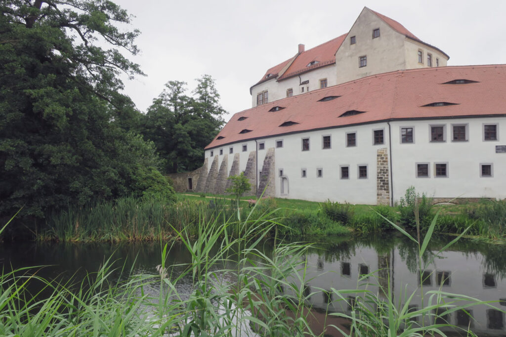 Blick auf ein historisches Schlossgebäude mit weißen Wänden und rotem Satteldach. Die linke Seite des Gebäudes wird von einem kleinen Teich oder Wassergraben gesäumt, in dem sich die Fassade spiegelt. Vordergrund: hohe grüne Schilfpflanzen am Ufer. Links ist ein dichter, hoher Baum zu sehen, der das Schloss teilweise einrahmt. Das Schloss wirkt solide, mit zahlreichen kleinen rechteckigen Fenstern, die sich gleichmäßig über die Fassade verteilen. Die Szenerie ist ruhig, leicht bewölkter Himmel im Hintergrund.