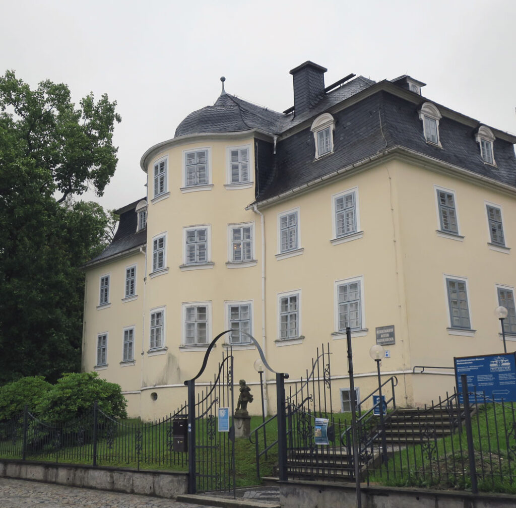 Ein zweistöckiges, cremefarbenes historisches Gebäude mit Schieferdach steht auf einer Grünfläche. Die symmetrische Fassade zeigt weiße Fensterrahmen und graue Läden; links ein runder Erker mit Kuppel, rechts ein Schornstein. Vor dem Eingang führen ein schmiedeeisernes Tor und eine Steintreppe hinauf, daneben steht eine kleine Bronzestatue. Ein Schild weist auf das „Museum Mannhardt“ hin. Der bedeckte Himmel verleiht der Szene eine ruhige, gedämpfte Stimmung.
